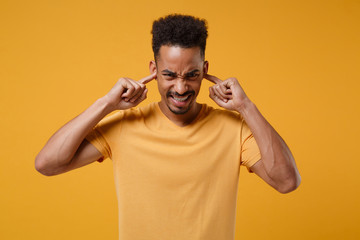 Displeased young african american guy in casual t-shirt posing isolated on yellow orange background, studio portrait. People emotions lifestyle concept. Mock up copy space. Covering ears with fingers.