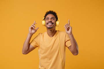 Funny young african american guy in t-shirt posing isolated on yellow orange background. People lifestyle concept. Mock up copy space. Listen music with headphones dancing pointing index fingers up.