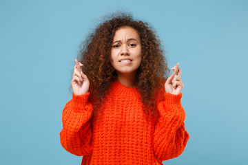 Young african american girl in casual orange knitted clothes keep fingers crossed eyes closed...