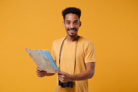 Smiling young african american guy in casual t-shirt with retro vintage photo camera posing isolated on yellow orange background. People lifestyle concept. Mock up copy space. Hold in hands city map.