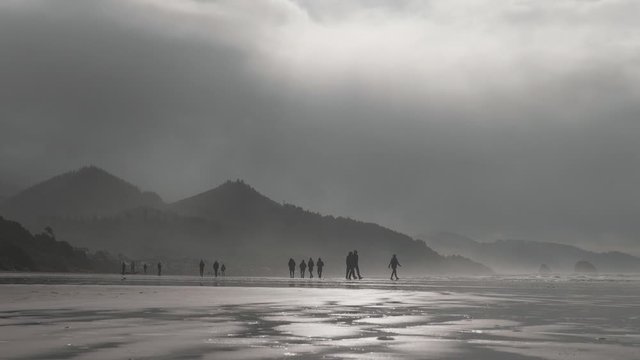 People walking across sandy beach at Cannon Beach Oregon on a moody overcast cloudy day in mist with mountains in background slow motion 