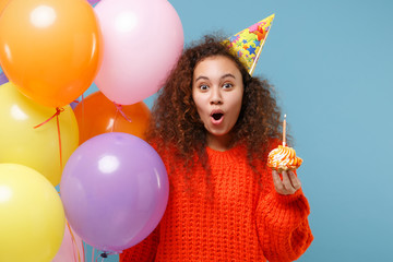 Shocked young african american girl in casual orange knitted clothes, birthday hat isolated on pastel blue background. Holiday party concept. Celebrating hold colorful air balloons cake with candle.