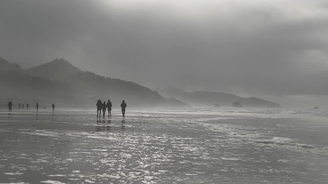 People walking on sandy beach at Cannon Beach Oregon on a moody overcast cloudy day in mist with mountains in background slow motion 