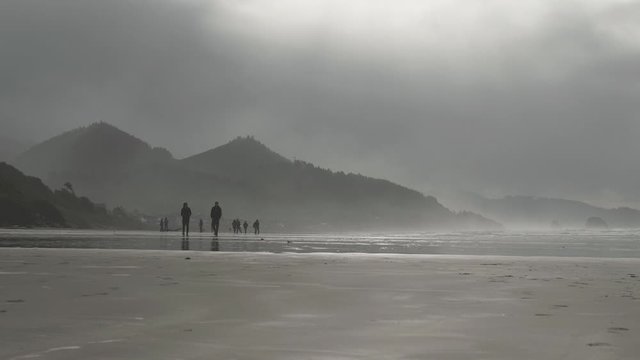 People walking across sand beach at Cannon Beach Oregon on a moody overcast cloudy day in mist with mountains in background slow motion 