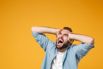 Mad young bearded man in casual blue shirt posing isolated on yellow orange wall background studio portrait. People lifestyle concept. Mock up copy space. Screaming, looking up, putting hands on head.