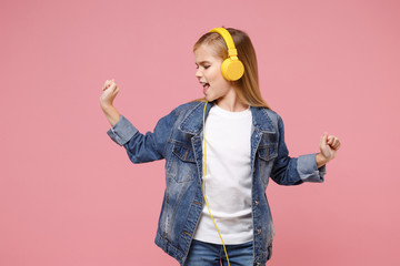 Funny little kid girl 12-13 years old in denim jacket isolated on pastel pink background in studio. Childhood lifestyle concept. Mock up copy space. Listen music with headphones dancing sing song.