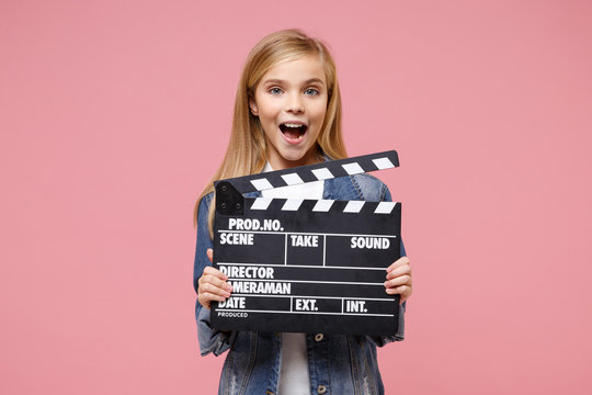 Excited little blonde kid girl 12-13 years old in denim jacket posing isolated on pastel pink background. Childhood lifestyle concept. Mock up copy space. Hold classic black film making clapperboard.