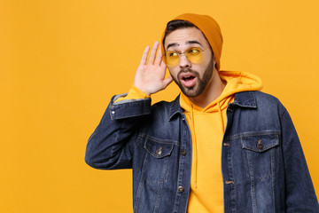 Curious young hipster guy in fashion jeans denim clothes posing isolated on yellow orange background studio portrait. People lifestyle concept. Mock up copy space. Try to hear you with hand near ear.