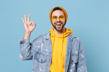 Naklejka premium Funny young hipster guy in fashion jeans denim clothes posing isolated on pastel blue background studio portrait. People sincere emotions lifestyle concept. Mock up copy space. Showing OK gesture.