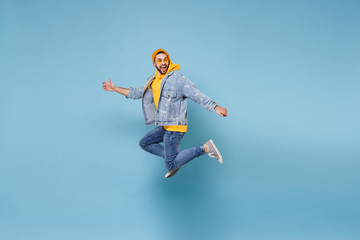 Side view of funny young hipster guy in fashion jeans denim clothes posing isolated on pastel blue background. People lifestyle concept. Mock up copy space. Jumping, spreading hands, looking aside.