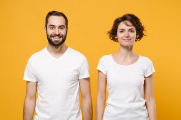 Fototapeta premium Smiling young couple friends bearded guy girl in white blank empty t-shirts posing isolated on yellow orange background studio portrait. People lifestyle concept. Mock up copy space. Looking camera.