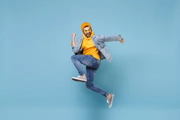 Fotobehang Side view of happy young hipster guy in fashion jeans denim clothes posing isolated on pastel blue background studio portrait. People lifestyle concept. Mock up copy space. Jump doing winner gesture. © ViDi Studio