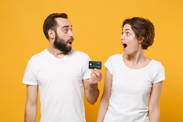 Shocked couple friends bearded guy girl in white blank empty t-shirts isolated on yellow orange background. People lifestyle concept. Mock up copy space. Hold credit bank card, looking at each other.