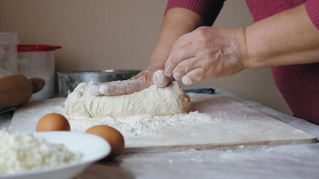 Close-up hands of senior female is kneading a dough in flour on the table at home kitchen, side view. Mature woman put a sticky dough in flour on the table and start kneading it. 