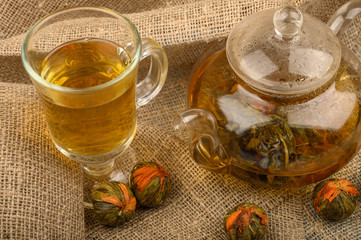 Flower tea brewed in a glass teapot, a glass of tea and balls of flower tea on a background of rough homespun fabric. Close up.