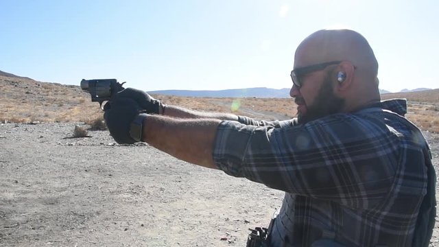 Bald man with beard shooting a revolver at an outdoor desert firing range. 24fps.