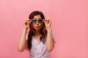 Cute lovely young woman in round sunglasses standing and sending kiss over pink background