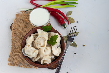 Boiled dumplings with feathers of green onions. In the background are greens, red peppers and bay leaves. In a transparent bowl sour cream.