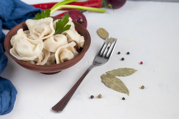 Boiled dumplings with feathers of green onions. In the background are greens, red peppers and bay leaves. In a transparent bowl sour cream.