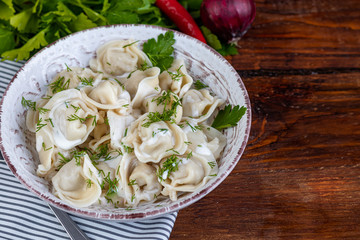 Boiled dumplings with feathers of green onions. In the background are greens, red peppers and bay leaves. In a transparent bowl sour cream.