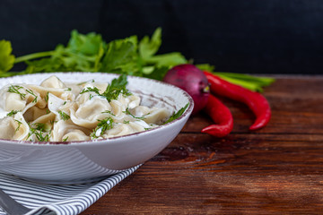Boiled dumplings with feathers of green onions. In the background are greens, red peppers and bay leaves. In a transparent bowl sour cream.