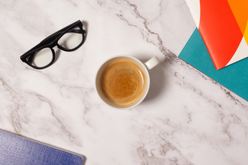 White cup of coffee on marble background with colorful notebooks and classical glasses 