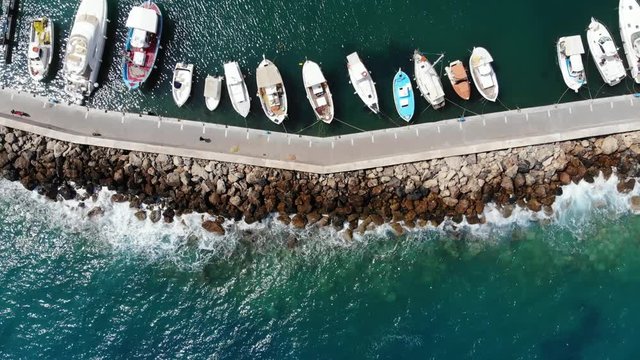 Aerial shot of sea wall at small Cretan port, many boats at quiet shelter, waves splash on rough stones from other side. Camera slowly move up, reveal wider view of breakwater
