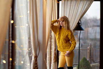 Portrait of a young pretty redhead woman wearing yellow sweater and yellow pants standing near window and looking away.