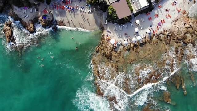 Yin and yang shaped beach, half with clear water and other hard shallow rock. People rest on small sand area, many sun loungers around. Top-down aerial shot. Sea waves wash stones