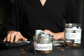 Woman using calculator near glass jar with money and tag BUSINESS on black table, closeup