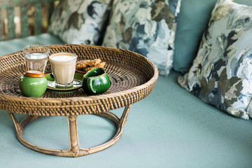 Rattan tray with cappuccino, milk gravy boat, sugar bowl, glass of water and cookies on blue sofa with pillows. Lazy morning concept.