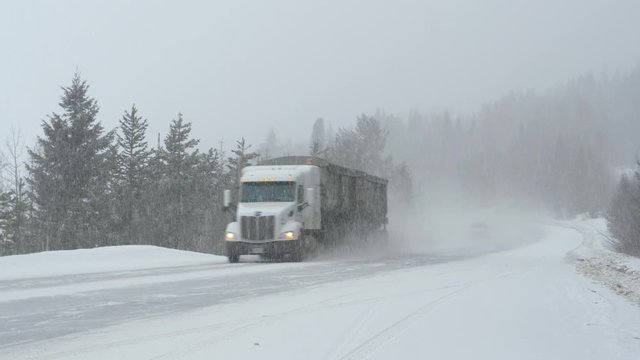 Freight truck leads the way for other vehicles driving through a blizzard covering rural Canada with fresh snow. Cars and truck carefully moves along an interstate road during a punishing snowstorm.