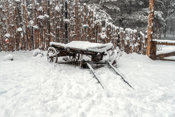 Tree branches in the winter forest under the snow, a wooden fence and a cart.
