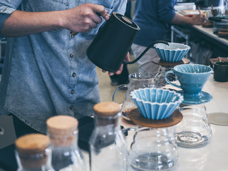 Barista in blue shirt pouring hot water from black kettle into coffee ground with filter on white counter bar.