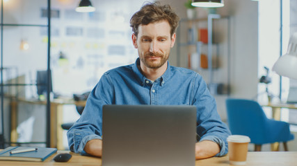 Young Handsome Man Works on a Laptop Computer in Cool Creative Agency in a Loft Office. He has a Take-away Coffee and a Notebook on the Table. He Wears a Jeans Shirt.
