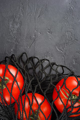 Red tomatoes in black net bag on gray concrete background. Healthy food, eco, diet concept. Top view, flat lay, macro, copy space, vertical format