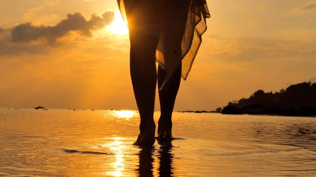 Close Up At  Foot Of Young Woman Walking In At Seashore  Wearing A Long Flowing Dress As Sunset Time In The Distance