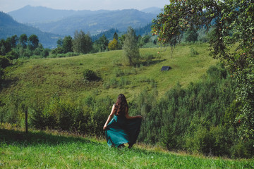 Happy, brunette girl running on the lawn. Carpathians mountains at the background. Young romantic woman in long, silk, green dress on the mountainside. Style of the goddess.