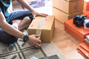 Thai young male online seller and start up small business owner sealing a box with tape and packing parcels for delivery at home.