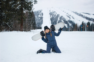Fototapeta na wymiar Beautiful young woman with the braids in grey winter costume is on her knees smiles and holds in the hands snowboard on a background of ski resort. Sunny winter day