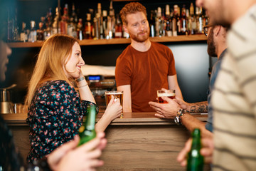 Fototapeta premium Cheerful friends leaning on bar counter, drinking beer and chatting with bartender. Night out.