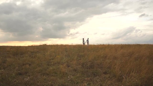 A happy couple of lovers slowly walks through a dry field in a strong wind, a man leads a girl by the hand. Wide shot. Happy young man leads his pregnant wife by the hand in the field at sunset.