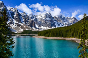 Fototapeta na wymiar This pristine Moraine Lake overlooks the icy rocky mountains and pine forest. The light breeze gently ripples the turquoise water towards the rocky edge on this partially cloudy day.