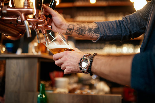 Tattooed caucasian barman pouring beer while standing in pub. Selective focus on hand.