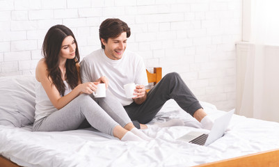 Young couple enjoying coffee and browsing on laptop in bed