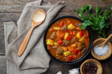 Goulash, beef stew or bogrash soup with meat, vegetables and spices in cast iron pan on wooden table. Hungarian cuisine. Rustic style. Top view. - obrazy, fototapety, plakaty