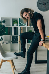 fashionable casual businessman in total black outfit posing in office on table
