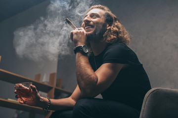 Low angle view of smiling fashionable businessman in black outfit smoking cigar and drinking whiskey