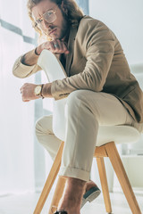 elegant fashionable businessman posing on chair and looking at camera in sunlight in office