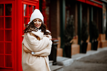 Street winter portrait of a young smiling woman in a white hat. A model poses against the backdrop of the city's Christmas decorations.